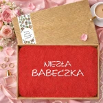 Czerwony ręcznik z haftem „Niezła Babeczka” ułożony w otwartym pudełku na różowym tle