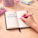 Otwarty notes w kratkę na biurku, dłoń zapisuje stronę różowym długopisem