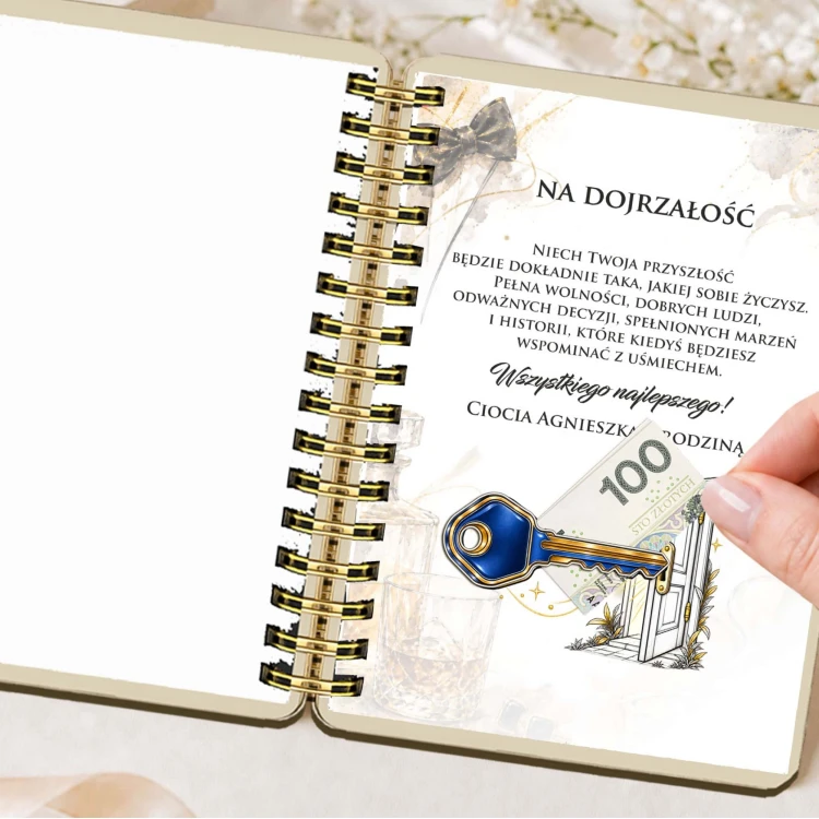Otwarty album na pieniądze z widoczną kartką zawierającą drukowane życzenia, ilustrację klucza i drzwi oraz miejsce na umieszczenie banknotu, trzymane przez dłoń