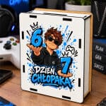 Drewniana skarbonka z kolorowym nadrukiem przedstawiającym postać chłopaka w okularach trzymającego liczby oraz napis, umieszczona na biurku obok kontrolera do gier